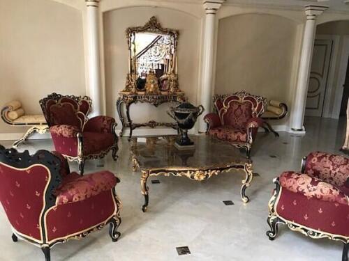 custom luxury living room furniture consisting of 4 wingback arm chairs with burgundy brocade cushions. matching marble topped coffee table, mirror, and console table feature the same black and gold woodwork as the armchairs. By Nino Madia.