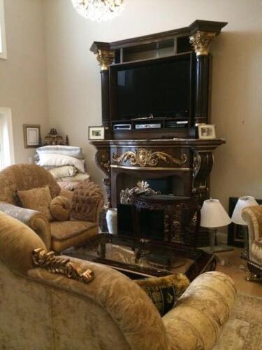 custom fireplace surround and TV entertainment center stacked on the mantle featuring columns, and gilded decorative accents. plush living room set with sofa and two arm chairs with glass inlay coffee table and matching wood console table.